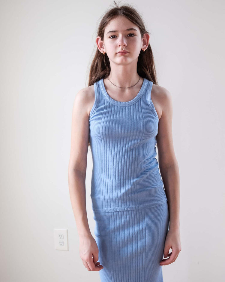 A young person with long brown hair stands against a white wall, wearing the sunchild fernando tank in mare, a sleeveless, ribbed light blue dress made from 100% cotton. They face the camera with a neutral expression; an outlet is visible behind them.