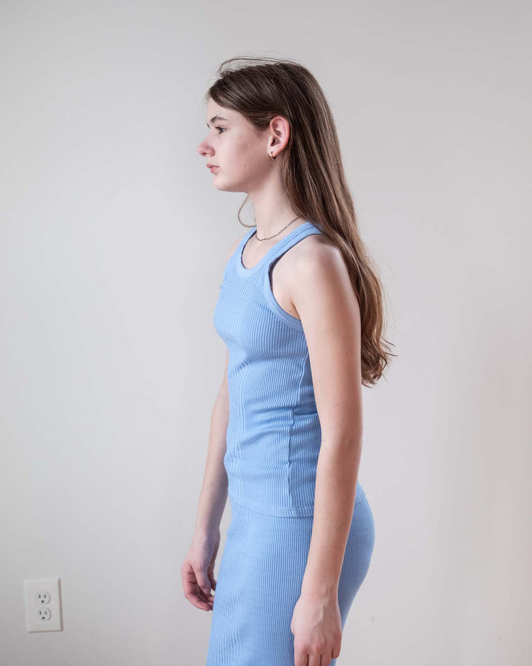 A young woman with long brown hair stands in profile against a light wall, wearing the fitted sunchild fernando tank in mare and a matching skirt, both 100% cotton. A white electrical outlet appears on the wall.