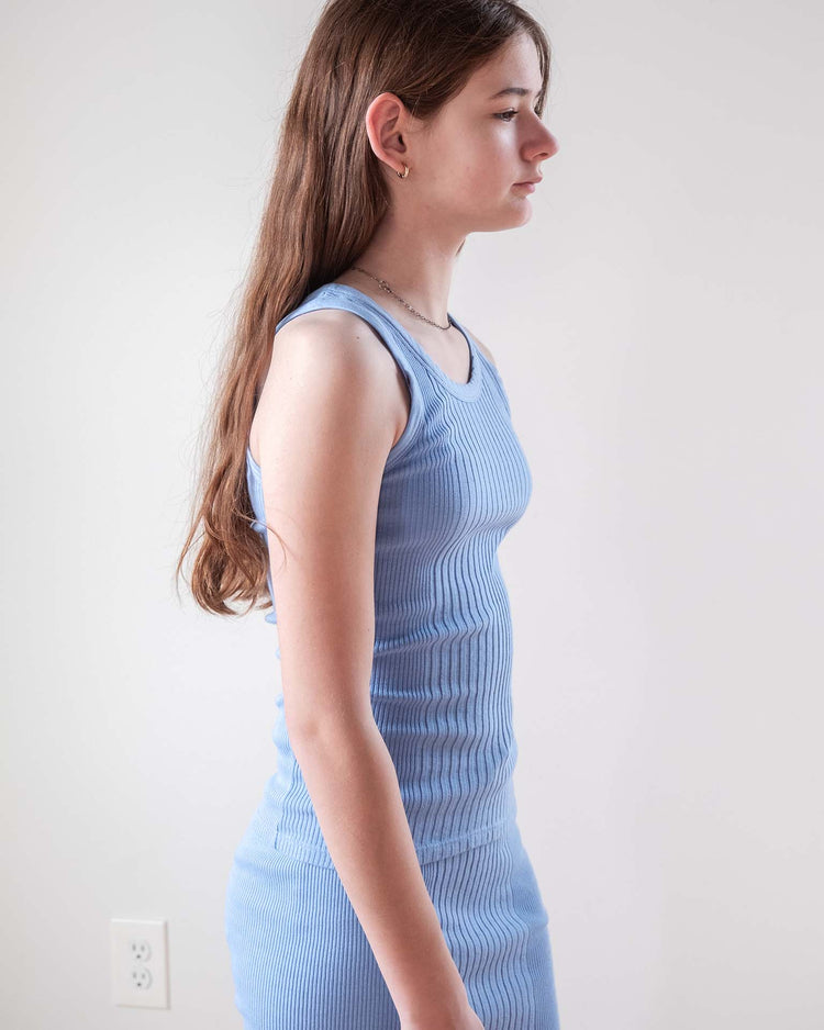 A young person with long brown hair stands in profile against a white wall, wearing the sleeveless, ribbed fernando tank in mare by sunchild, made from 100% cotton.