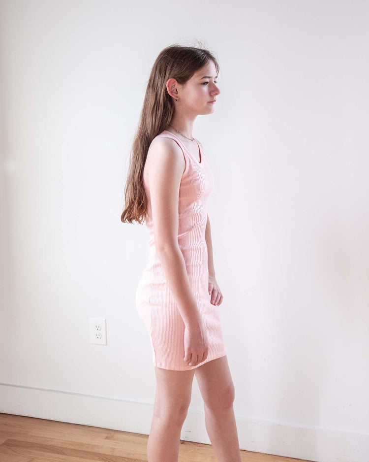 A young woman with long brown hair stands in profile against a plain white wall, wearing the Sunchild pipa dress in baby. The ribbed, fitted tank dress is light pink 100% cotton. A wooden floor and a white electrical outlet are behind her.