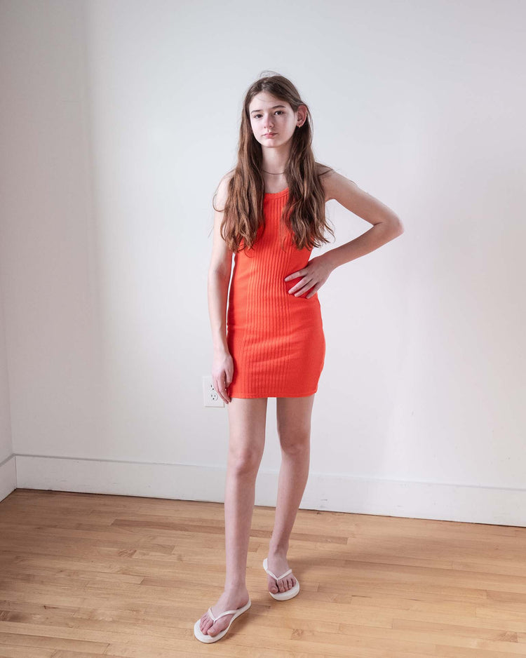 A young person with long brown hair stands indoors against a white wall on a wooden floor, wearing the Sunchild pipa dress in tangerine and white flip-flops. One hand is on their hip, and they have a neutral expression.