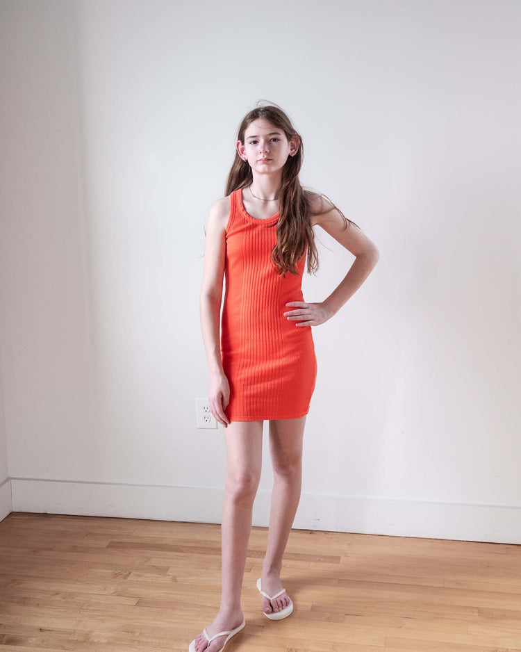 A young person with long brown hair stands indoors against a white wall, wearing the sunchild pipa dress in tangerine and white sandals, with one hand on their hip.