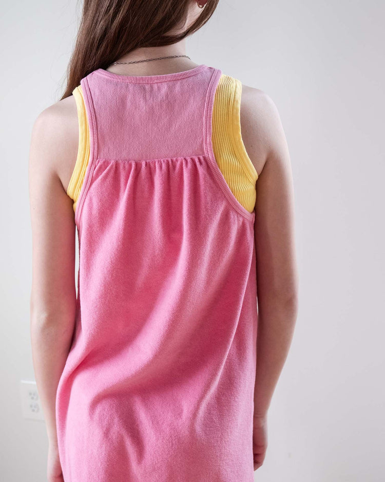 A girl with long brown hair stands facing away, wearing the sunchild bilbao dress in glamour—a 100% cotton pink tank dress with yellow trim—against a plain light background.