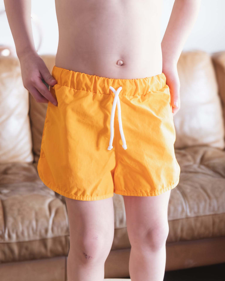 A child wearing bright orange Bahia Board Shorts in Inca by Sunchild stands indoors in front of a tan leather couch, hands at their sides and not wearing a shirt.