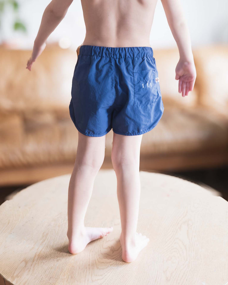 A child, seen from behind and barefoot on a wooden table, wears only sunchild bahia board shorts in obscur with arms gently extended. A blurred brown sofa appears in the background.