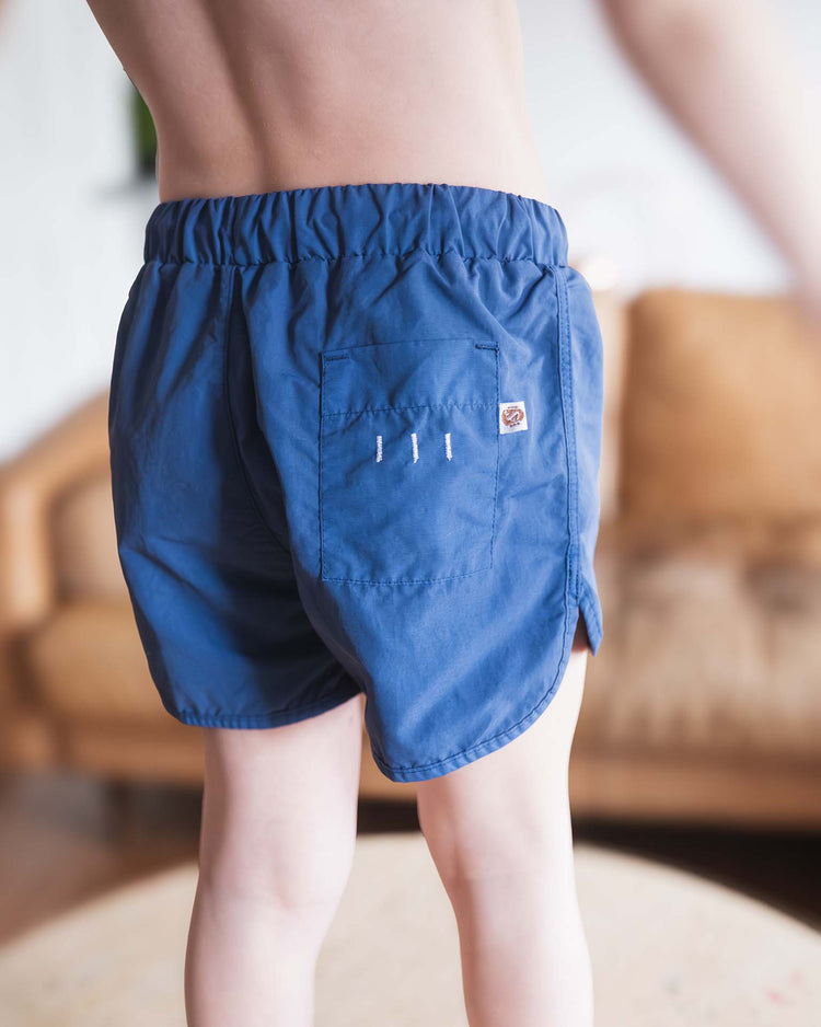 A child stands indoors, seen from behind, wearing Sunchild bahia board shorts in obscur with an elastic waistband and a pocket. A softly blurred brown couch is visible in the background.