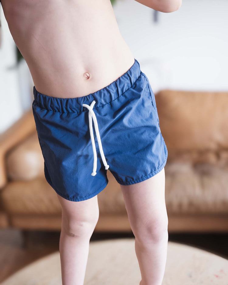 A child models the sunchild bahia board shorts in obscur, standing on a round wooden table with a tan couch blurred in the background; only their upper body and legs are visible, not their face.