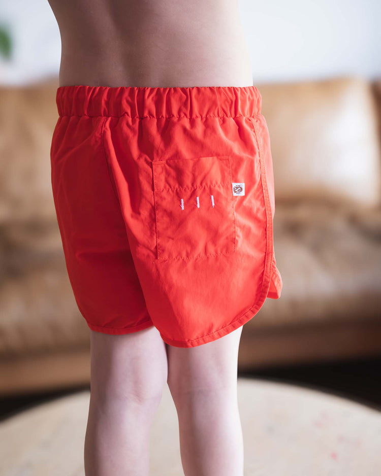 A child wears sunchild's bahia board shorts in tangerine, showing off the side and back pockets against an indoor background with a tan couch.