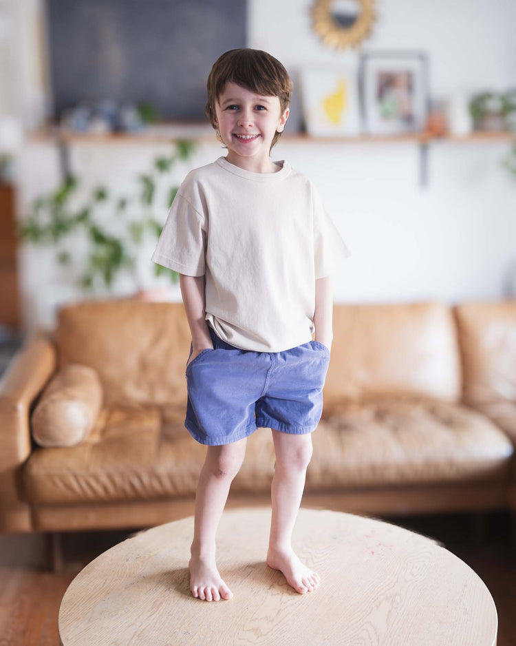 A young Sunchild with short brown hair wears sunchild's hunter shorts in obscur and a beige t-shirt, standing barefoot on a round wooden table in a cozy living room with a tan couch and plants in the background.