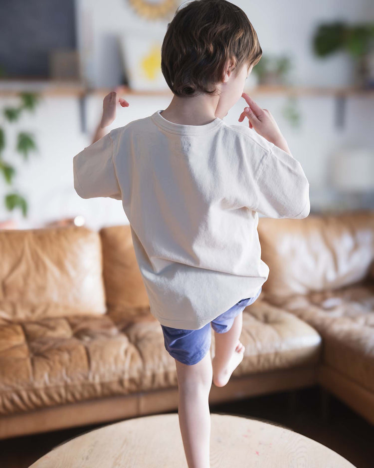 A child in Sunchild’s kazan tee in craie and blue shorts balances on one leg atop a wooden table, facing away, in a cozy living room with a brown couch and plants—a scene straight from the carefree world of Sunchild.