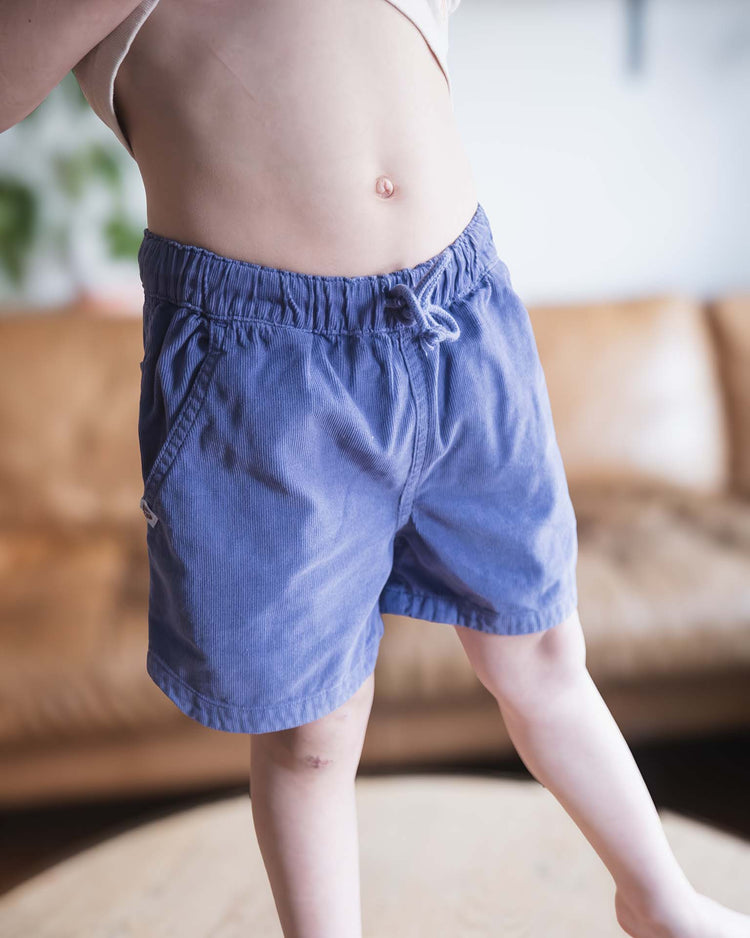 A young model from sunchild wears hunter shorts in obscur—a blue cotton pair with a drawstring waistband—and a white top, standing indoors with their midsection and legs visible. A brown couch is softly blurred in the background.
