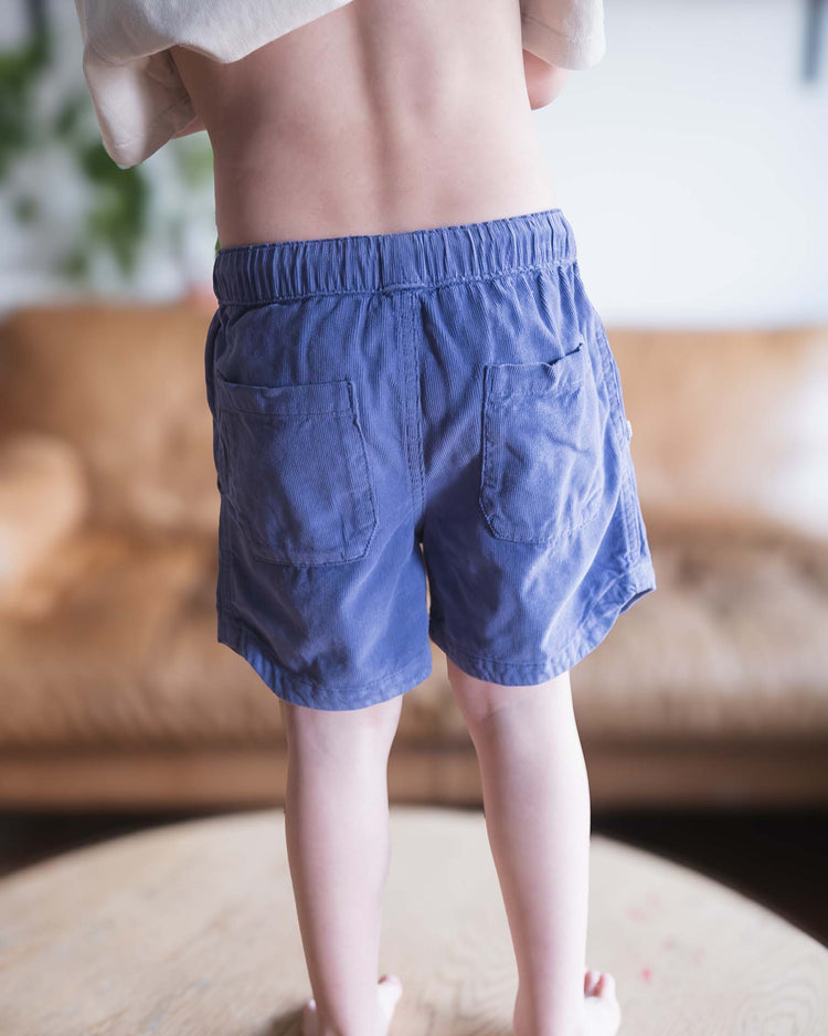 A Sunchild stands on a round wooden table, back to the camera, wearing sunchild hunter shorts in obscur with a drawstring waistband and a light shirt. A brown couch and green plants blur in the background.