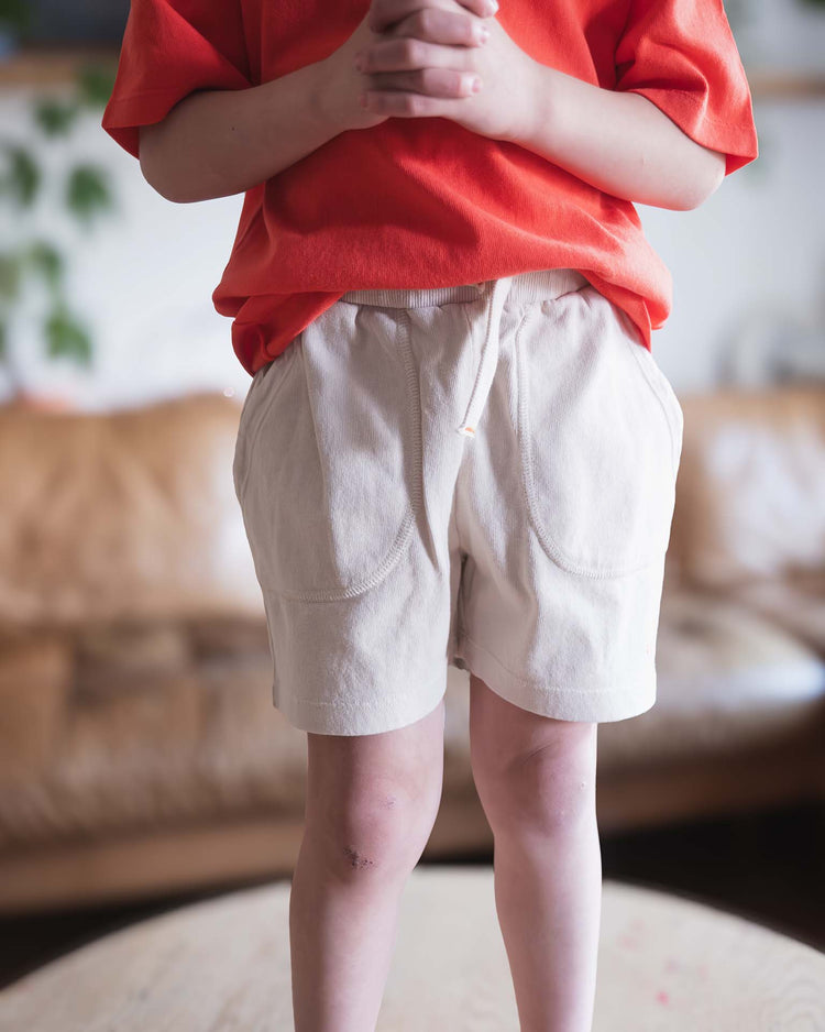 A Sunchild in a red shirt and merida shorts in craie by Sunchild, featuring a drawstring waist, stands indoors on wooden flooring with hands clasped. The blurred background shows a brown couch and green plants.