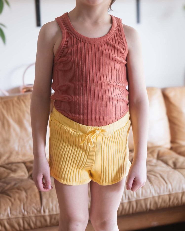 A child stands indoors in front of a tan leather couch, wearing a sunchild fernando tank in cacao. The child's face is out of frame.