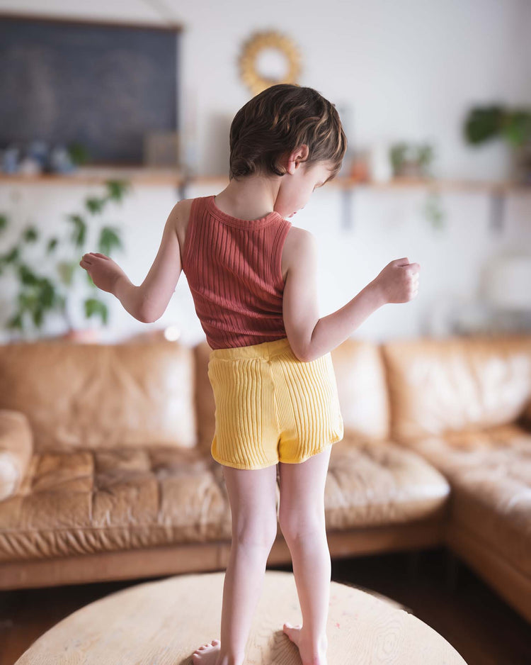 A young child stands on a round wooden table, wearing the Sunchild fernando tank in cacao and yellow shorts, with their back to the camera in a cozy living room with a tan sofa and green plants in the background.