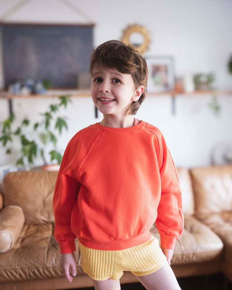 A young child with short brown hair smiles indoors, wearing the Sunchild douglas sweatshirt in tangerine and yellow shorts. A tan leather couch and green plants are visible in the background.