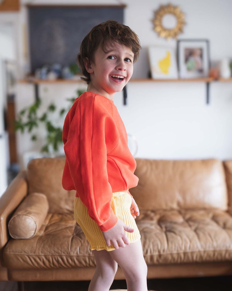 A young child wearing a Sunchild douglas sweatshirt in tangerine and yellow striped shorts smiles in front of a tan leather couch in a cozy, well-lit living room.