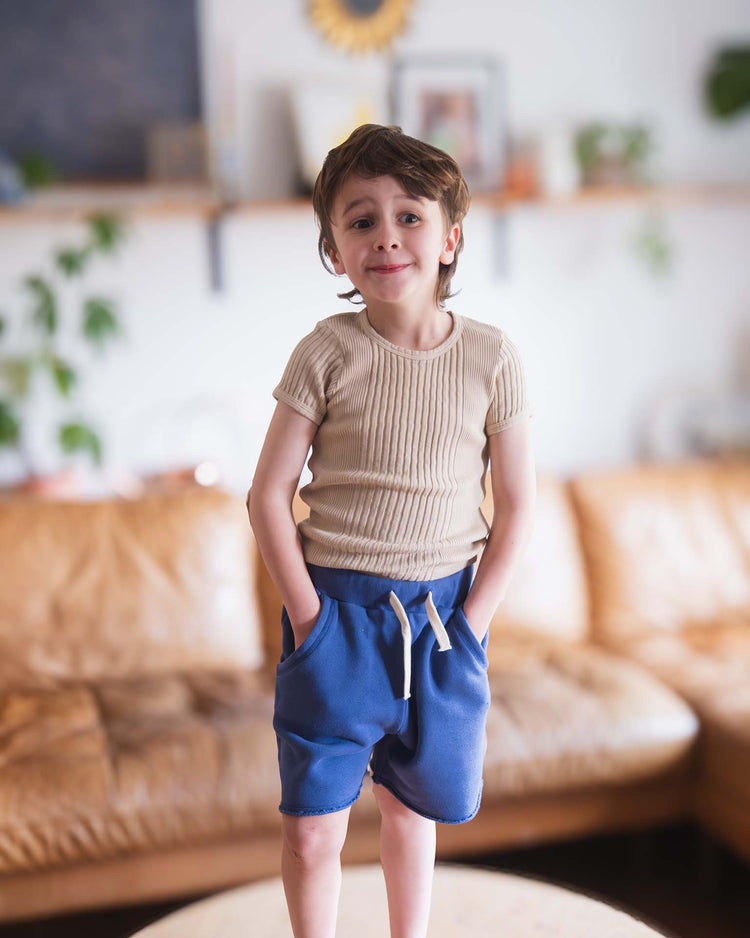 A young boy with short brown hair, wearing a beige ribbed shirt and Sunchild pico shorts in obscur with a drawstring waist, stands with his hands in his pockets on a table in a cozy living room with a tan leather couch and green plants.