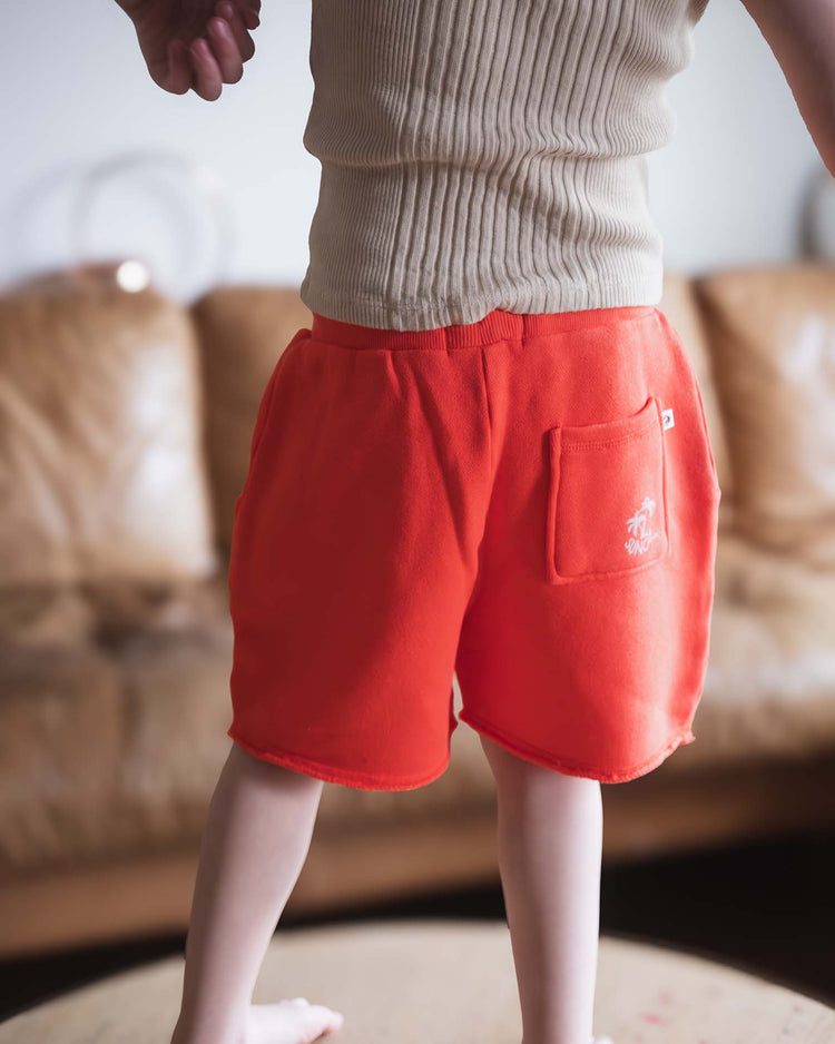 A child stands on a table in a beige ribbed tank top and Sunchild pico shorts in tangerine, featuring a drawstring waist and pocket. A blurred brown couch is visible in the background; the child's face is not shown.