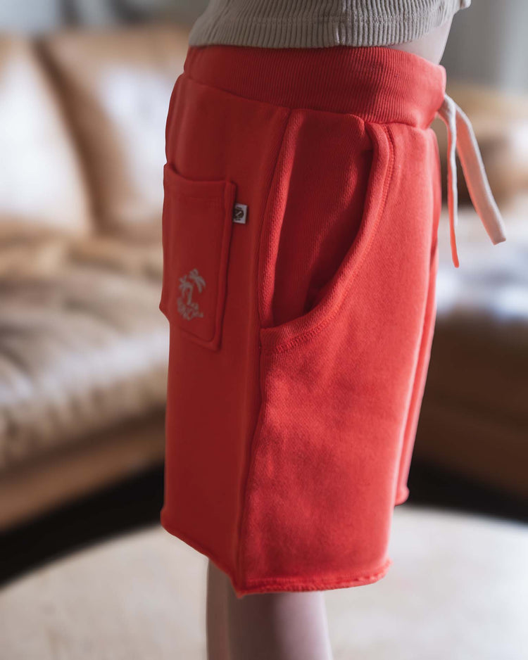 Someone wears Sunchild pico shorts in tangerine—a bright orange cotton short featuring a small side pocket and drawstring waist—while standing indoors with a blurred beige sofa in the background.
