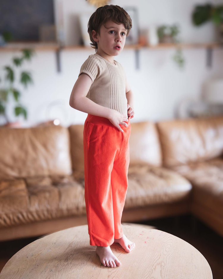 A young child with short brown hair stands barefoot on a round wooden table, wearing a beige shirt and sunchild's bluff pants in tangerine. A brown leather sofa and green plants appear softly blurred in the background.