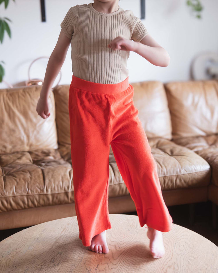A child stands barefoot on a wooden table, wearing a beige ribbed shirt and sunchild bluff pants in tangerine, with a tan leather sofa and plants in the background.