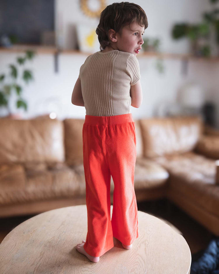 A young child with short brown hair stands barefoot on a round wooden table, wearing a beige ribbed shirt and sunchild’s bluff pants in tangerine. A brown sofa and green plants are visible in the background.