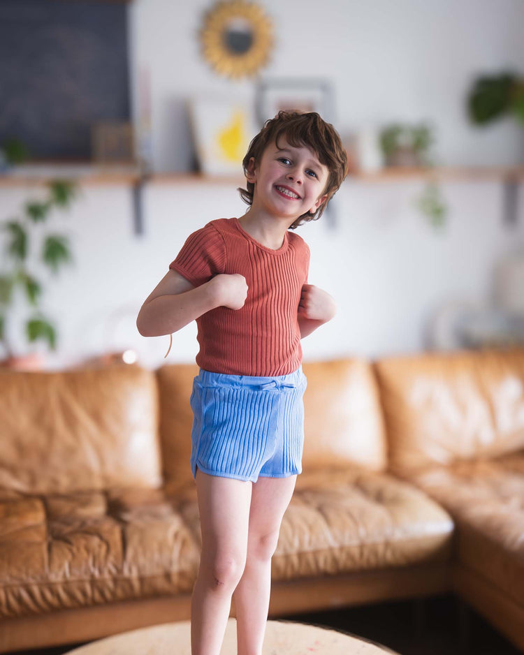 A young child with short brown hair smiles while wearing the sunchild loutro tee in cacao and light blue shorts, standing on a round table in a cozy living room with a brown leather sofa and plants in the background.