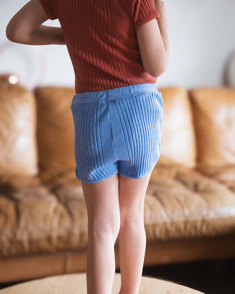 A child in a red ribbed shirt and Sunchild rio shorts in mare with a drawstring waist stands on a wooden surface, facing away from the camera. A brown leather couch is visible in the background.