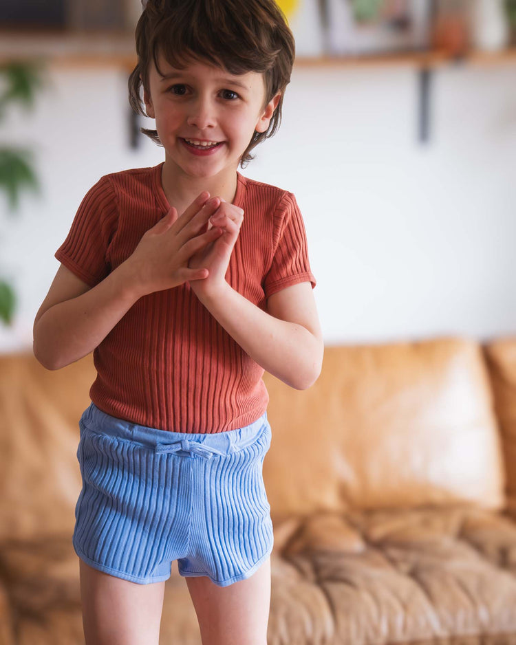 A young child with short brown hair smiles and clasps their hands, wearing a rust ribbed shirt and sunchild rio shorts in mare with a drawstring waist, standing before a tan couch in a cozy living room.