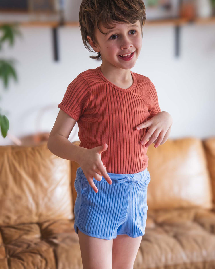 A smiling young child with short brown hair wears a sunchild loutro tee in cacao and blue shorts, standing indoors in front of a tan leather couch.