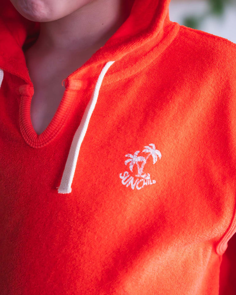 A person wearing a tangerine Sunchild sitges sleeveless sweatshirt featuring white drawstrings and an embroidered design of two palm trees above the word SUNCHILD.