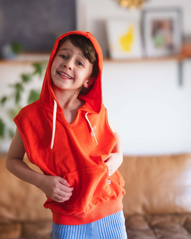 A smiling young child wears a Sunchild sitges sleeveless sweatshirt in tangerine, standing indoors and holding the hem. A softly blurred couch and wall decor are visible in the background.