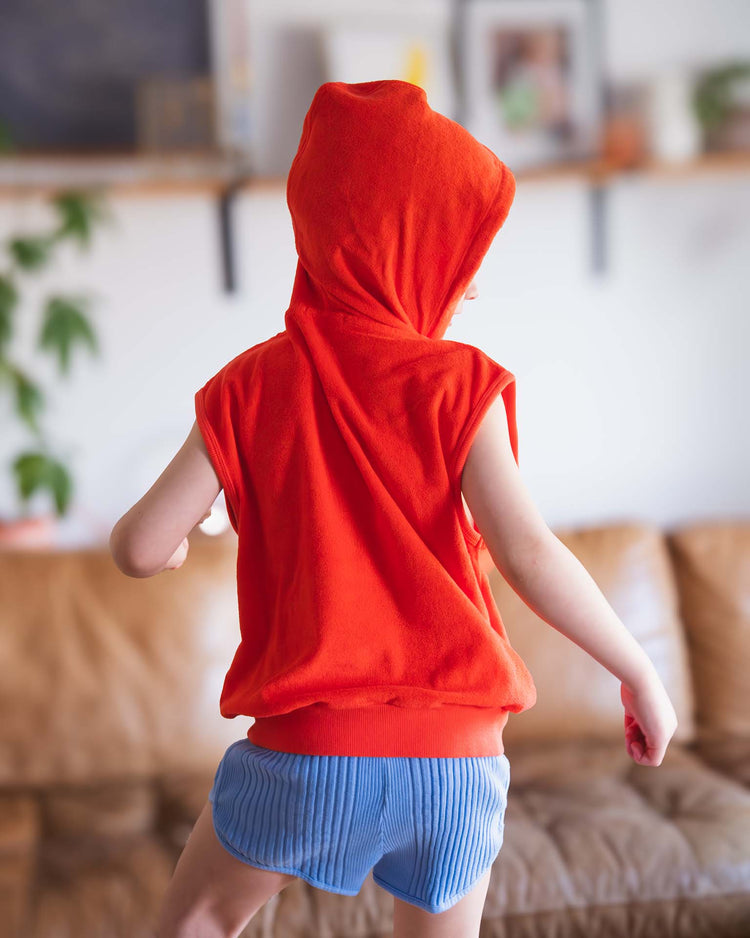 A child in a Sunchild Sitges sleeveless sweatshirt in tangerine and blue shorts stands with their back to the camera in a living room featuring a brown couch and a blurred background.
