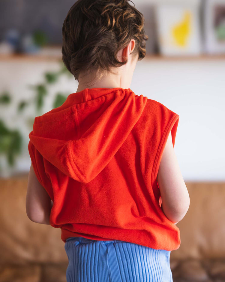 A child with short brown hair, seen from behind, wears the sitges sleeveless sweatshirt in tangerine by sunchild and light blue ribbed pants, standing indoors against a softly blurred background.