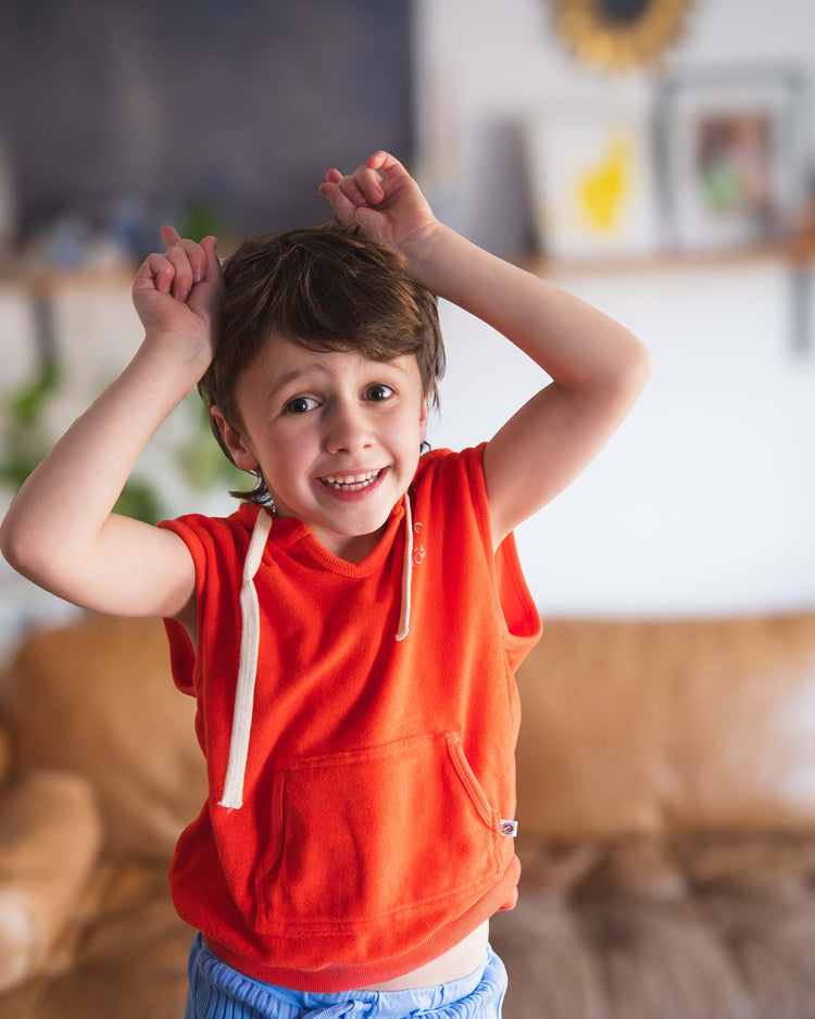 A young boy smiles indoors, wearing the sunchild sitges sleeveless sweatshirt in tangerine and blue shorts. He holds his hands up like animal ears; a blurred brown couch and shelves appear in the background.