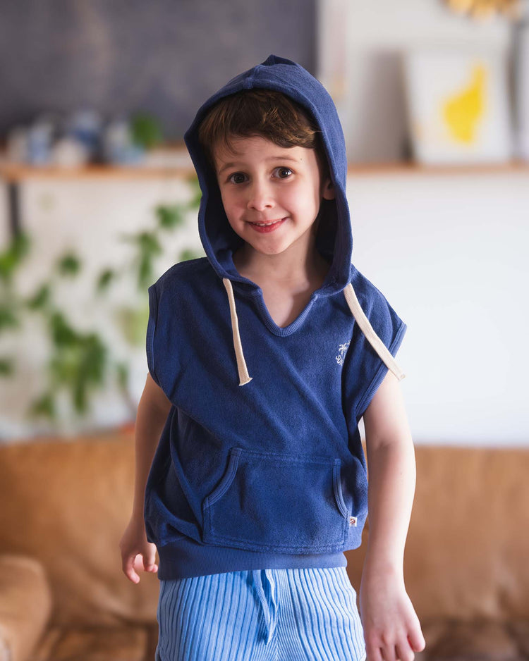 A young boy with short brown hair smiles indoors, wearing a Sunchild Sitges sleeveless sweatshirt in Obscur with the hood up and blue-and-white striped shorts. The softly blurred background features plants and shelves.