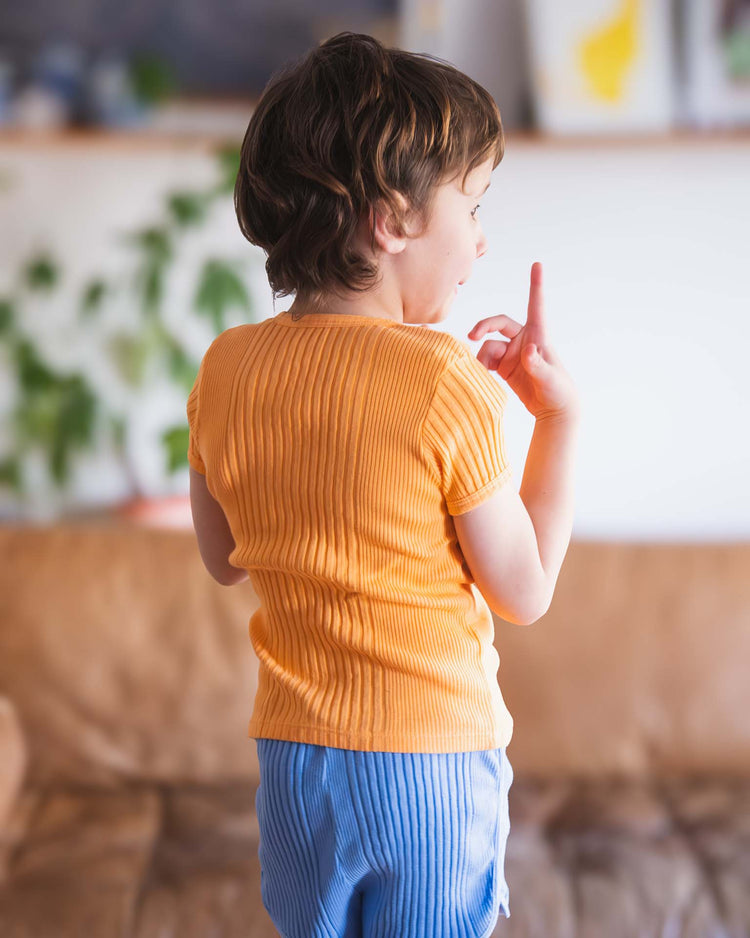 A young child in the sunchild loutro tee in inca stands indoors, facing away with their index finger raised. Softly blurred plants and shelves create a warm, charming atmosphere.