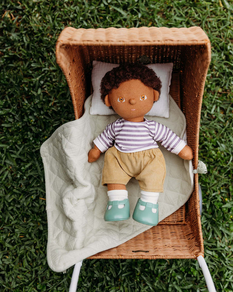 A Dinkum Dolls brown-skinned doll with curly hair, a striped shirt, tan shorts, and green shoes lies on a pillow, partly covered by the Strolley Bedding Set in Sage by Dinkum Dolls, in a wicker bassinet on grass.