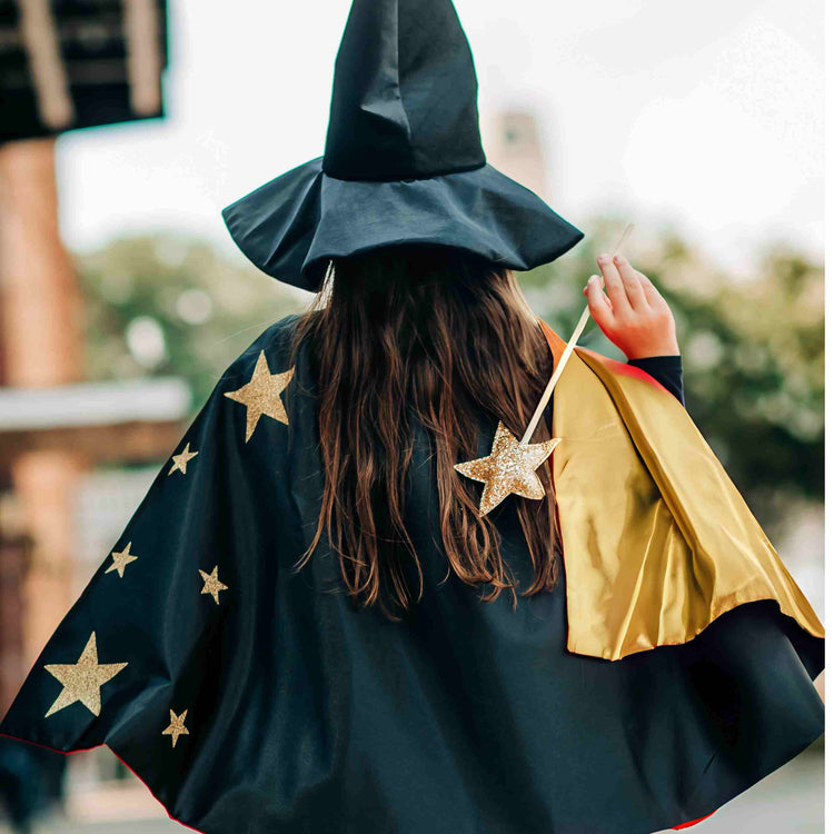 A child dressed as a wizard stands outdoors wearing Bailey and Ava's Spellbound Witch Costume Set in Black, featuring a glitter star cape, wand, and large velvet witch hat.