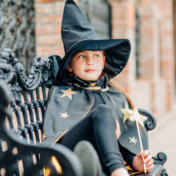 A young girl in the Bailey and Ava Spellbound Witch Costume Set in Black, complete with a glitter star cape and velvet hat, sits on an ornate bench holding a small wand.