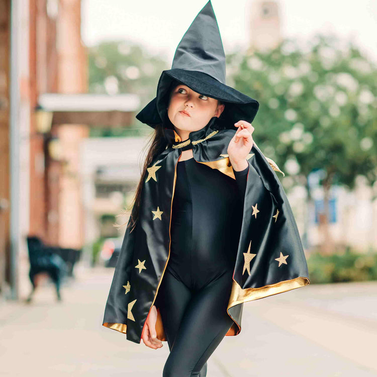 A child wearing the Spellbound Witch Costume Set in Black from Bailey and Ava stands outdoors, their outfit adorned with gold stars and completed by an enchanting velvet witch hat.