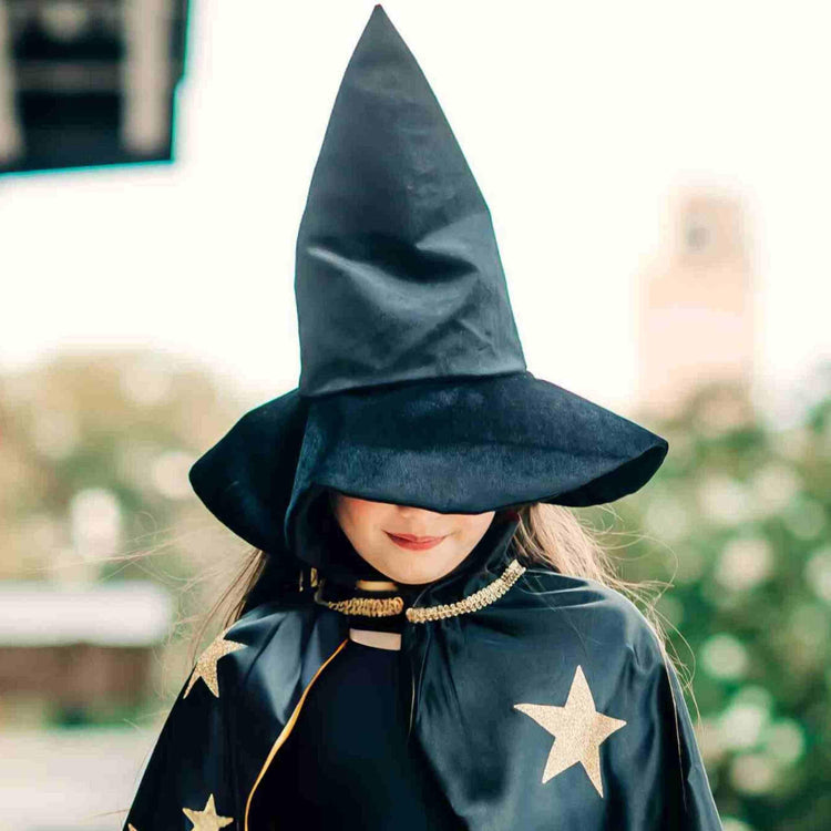 A child outdoors wears the Bailey and Ava Kids Velvet Witch Hat in black, its tall shape partially covering their face, along with a black cape decorated with gold stars.