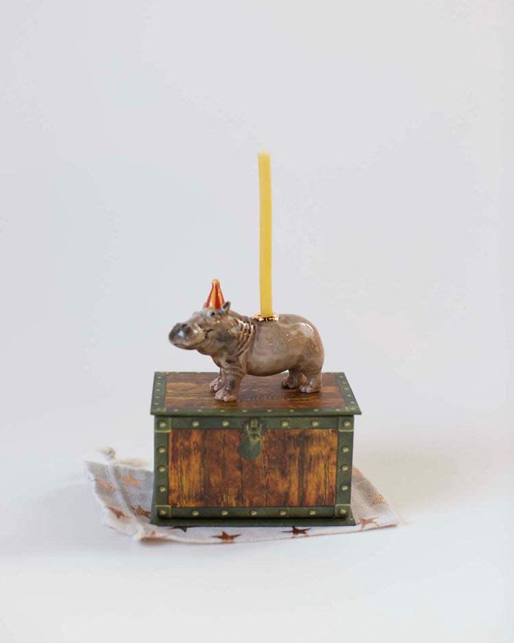 A hand-painted ceramic hippo cake topper from Camp Hollow on a small, decorative wooden chest with a yellow candle on its back, placed against a plain white background.