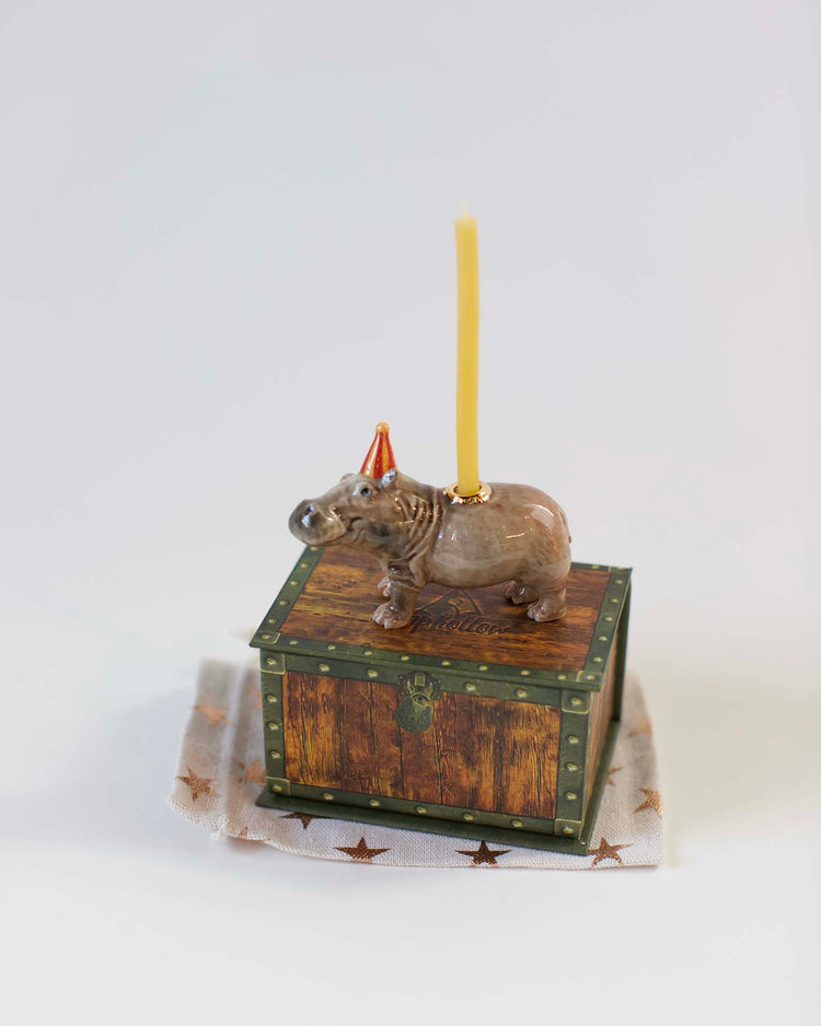 A decorative ceramic Camp Hollow hippo cake topper on top of an ornate wooden box, with a yellow candle fixed on its back, set against a plain white background.