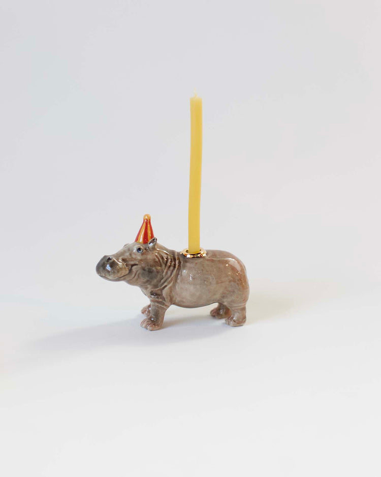 A hand-painted, ceramic camp hollow hippo cake topper with a yellow candle positioned on its back, set against a plain white background.