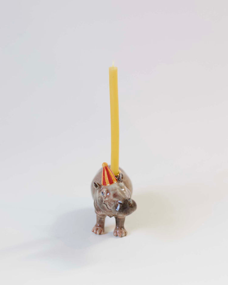 A hand-painted ceramic camp hollow hippo cake topper with a yellow candle positioned on its back, set against a plain white background.