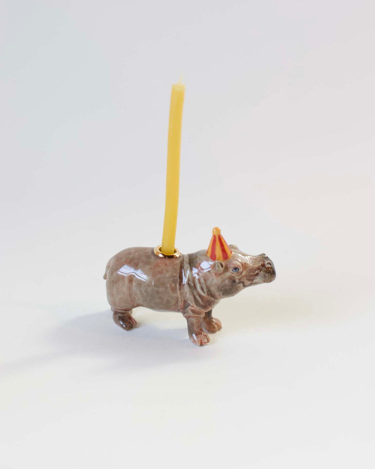 Camp Hollow hippo cake topper with a yellow candle mounted on its back, placed against a plain white background.