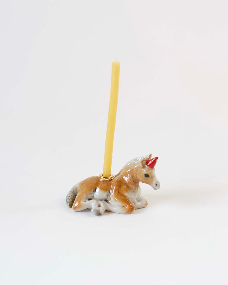 A hand-painted ceramic year of the horse cake topper with a yellow candle inserted, positioned against a plain white background. Made by Camp Hollow.