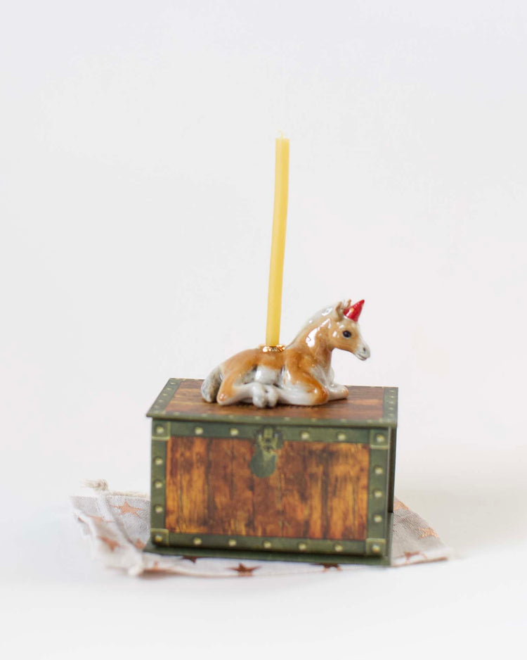 A Camp Hollow year of the horse cake topper with a single yellow hand-painted candle holder on its back, mounted on a small, decorative wooden chest with brass accents, set against a white background.